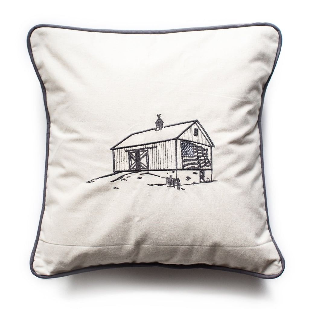 Ivory pillow with black ink and black edging. Image of a country barn with American flag on side and weathervane on top of roof.
