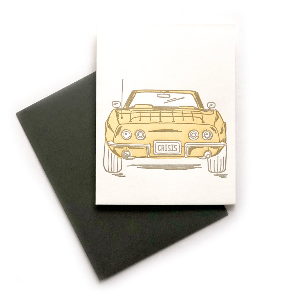 White card with image of a yellow convertible sports car with “CRISIS” in green text on the back license plate. A black envelope is included.