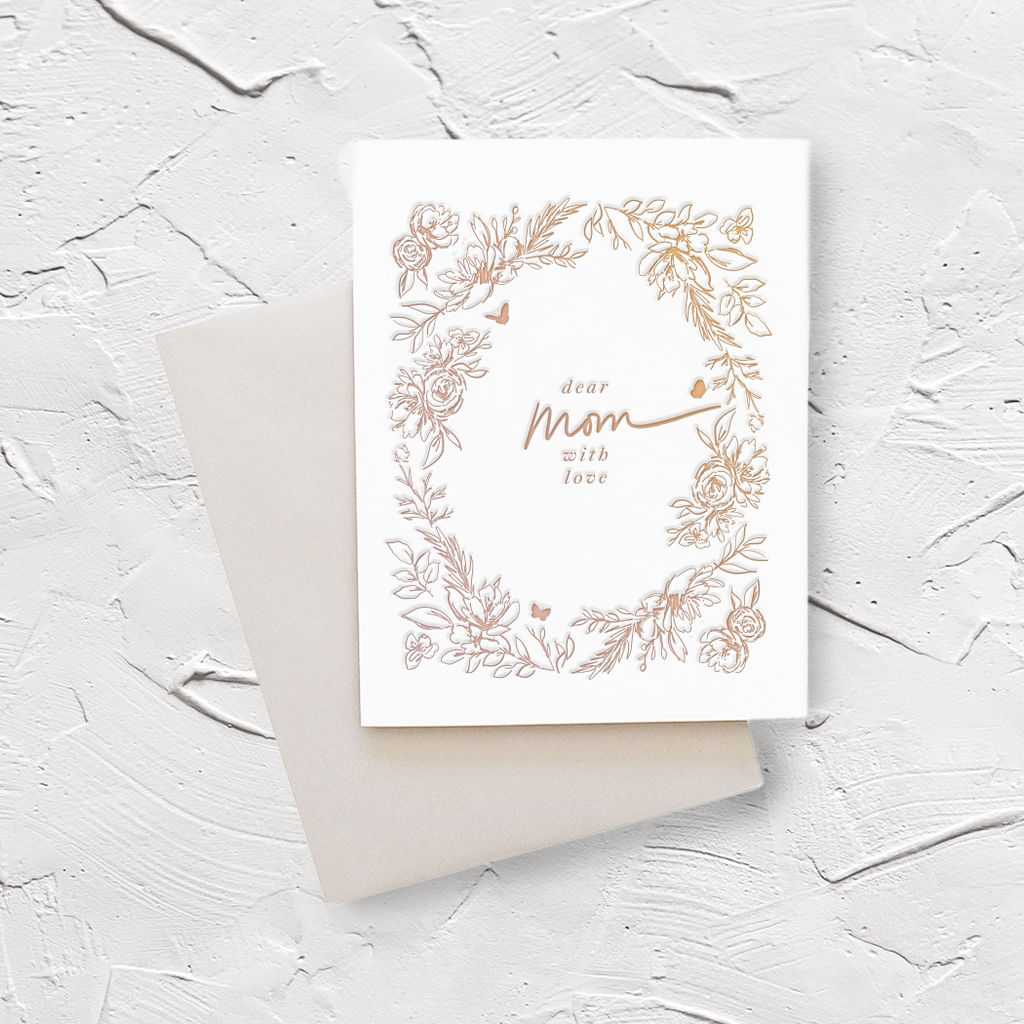 White card with gold text saying, “Dear Mom With Love”. Images of gold floral border. A gray envelope is included.