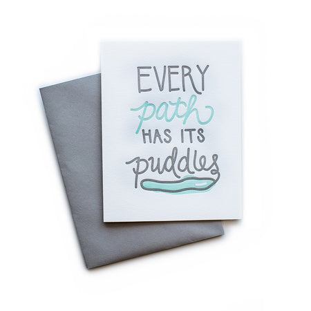 White card with gray and teal text saying, “Every Path Has Its Puddles”. Images of a puddle with blue water. A gray envelope is included.