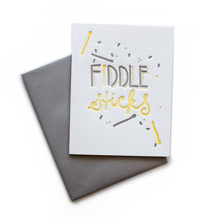 White card with gray and yellow text saying, “Fiddle Sticks”. Images of drumsticks and gray confetti. A gray envelope is included.