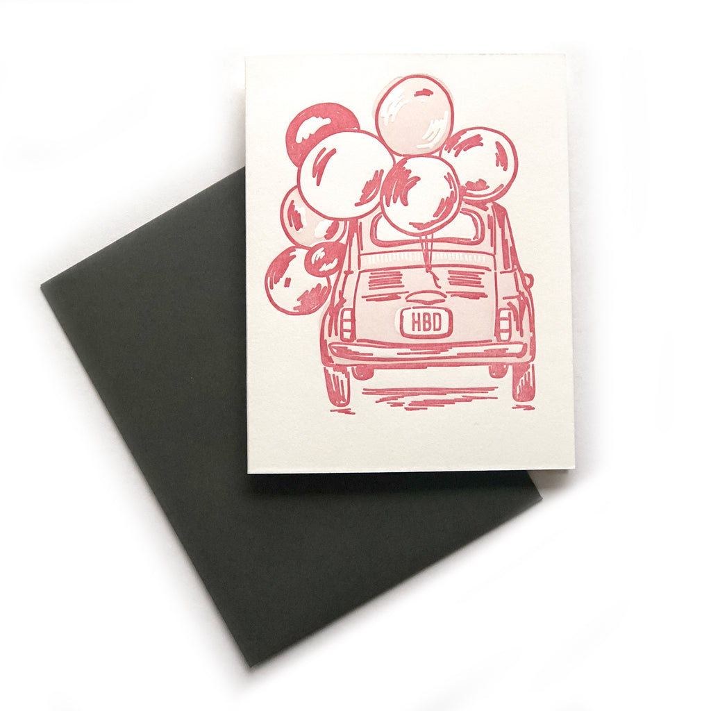 White card with image of a pink vintage beetle bug card with “HBD” on license plate. Red, white and pink balloons tied to back of car. A dark gray envelope is included.