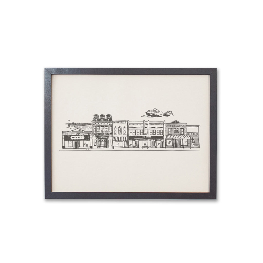 Art print with ivory background and black ink. Image of a hometown main street with buildings: market; fire station; Feed & Supply store; and dry goods store. Farmland in the distance and clouds in the sky.