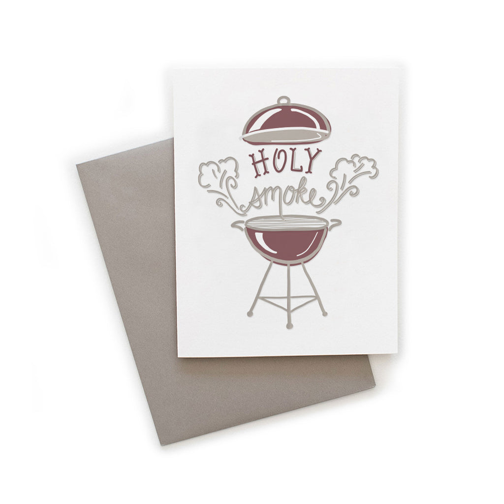 White card with gray and red text saying, “Holy Smoke”. Image of a classic bbq grill with smoke coming from each side. A gray envelope is included.