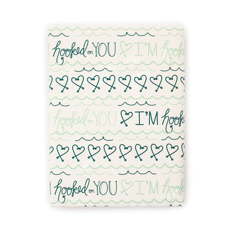 Ivory background with teal and navy blue text saying, “I’m Hooked On You”. Images of hearts made out of fishhooks and teal blue waves across paper.