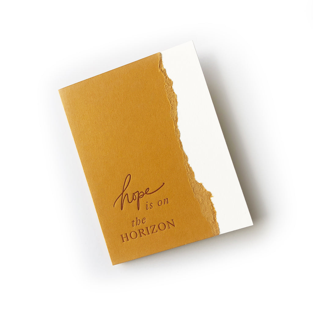 White and gold card with a curved torn edge where the two colors meet. Gold text saying, “Hope is on the Horizon”. A white envelope is included.