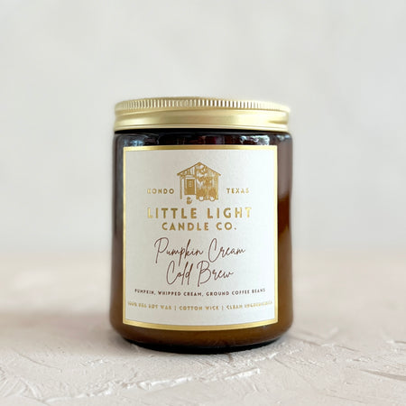 Brown glass jar with gold lid and white label with gold foil text saying, “Little Light Candle Co. Pumpkin Cream Cold Brew”. 