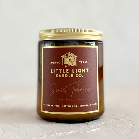Brown glass jar with gold lid and brown label with gold foil text saying, “Little Light Candle Co. Sweet Tobacco”.