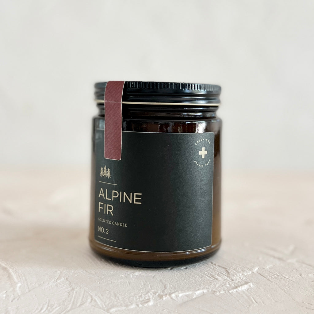 Brown jar with black lid and dark green label with white text saying, “Alpine Fir”. Images of 3 small fir trees.