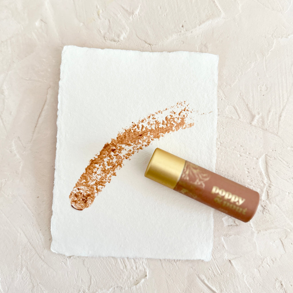 Orange colored tube with gold top and gold foil text saying, "Poppy & Pout Faye Lip Tint". Image of an outlined flower in gold foil.