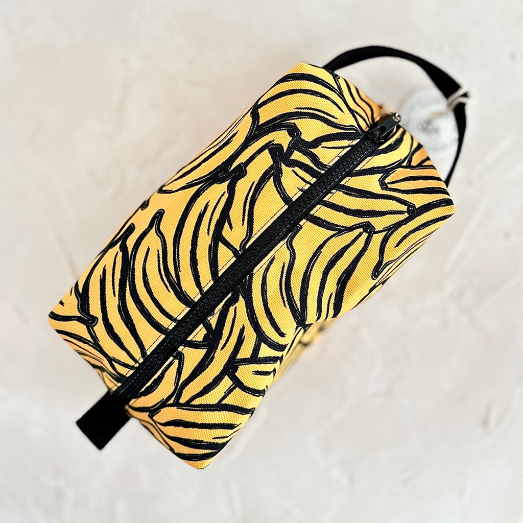 Rectangular travel pouch with yellow background and black bananas design. Black zipper across middle and black cloth handle on each end.