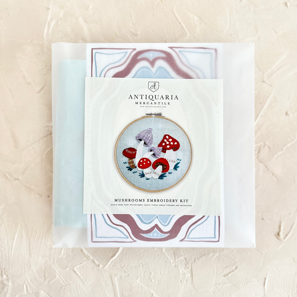 Ivory package with black text saying, “Antiquaria Mercantile Mushrooms Embroidery Kit”. Images of red and purple mushrooms with white stems and green leaves on ground.