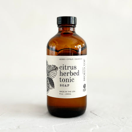 Brown bottle with black lid and white label with black text saying, “Citrus Herbed Tonic Soap”. Image of herb leaves.