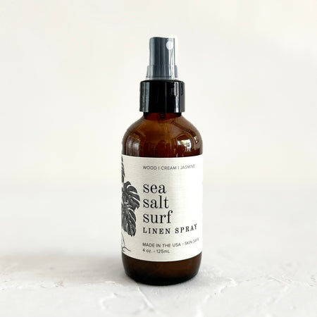 Brown bottle with black spray lid and white label with black text saying, “Sea Salt Surf Linen Spray”. Image of palm branch leaves.