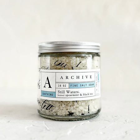 Glass jar with silver lid and white label with black text and blue accents. Text saying, “Archive Still Waters Fine Salt Soak”.
