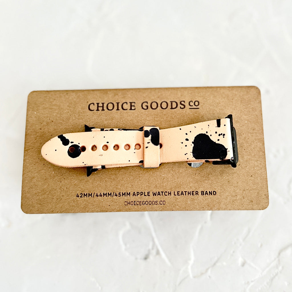White leather watch band with black paint splattered design. Packaged on a brown cardboard rectangle with black text saying, “Choice Goods Co.”