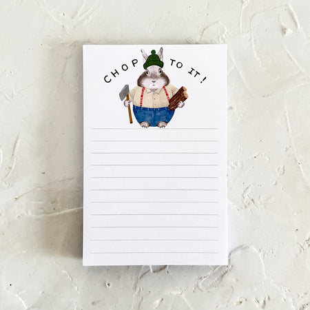 White notepad with image of a brown bunny wearing a white shirt, blue pants, red suspenders and a green knit hat holding an axe and logs of wood. Black text saying, 