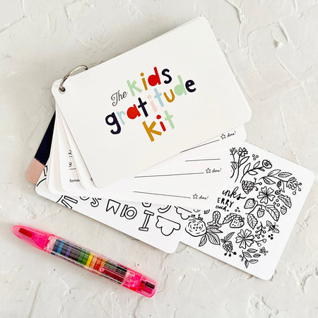 White rectangle cards on a silver ring. Cover has rainbow text saying, “The Kids Gratitude Kit”. Each individual card has black and white images for the kids to color.