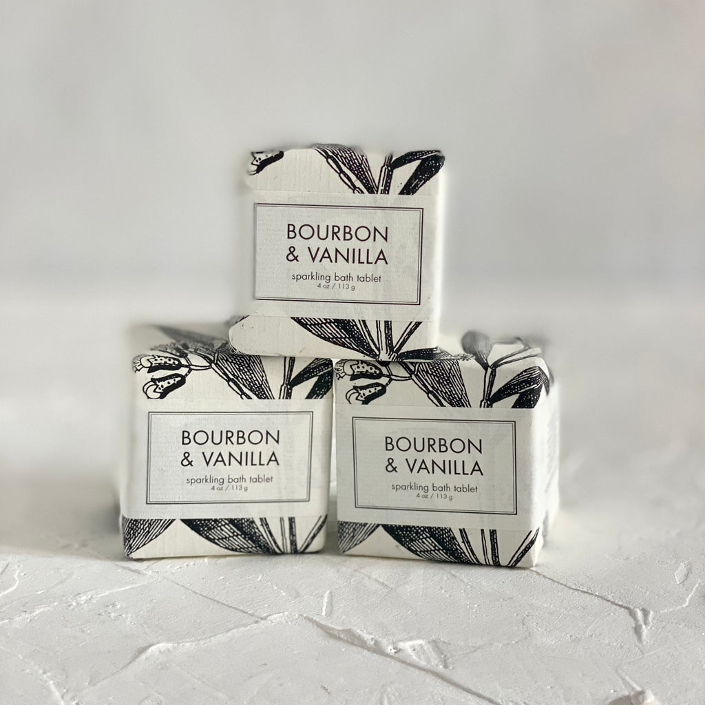 Packaged in a square white box with white label and black text saying, “Bourbon and Vanilla Sparkling Bath Cube”. Images of black vanilla bean plants.