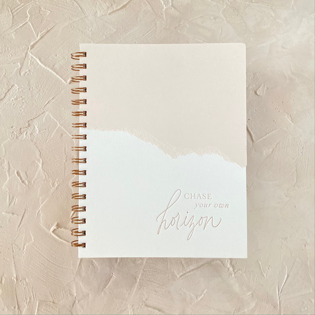 Ivory and tan cover with curved line where colors meet and text saying, “Chase Your Own Horizon”. Gold metal spiral on left side of notebook.