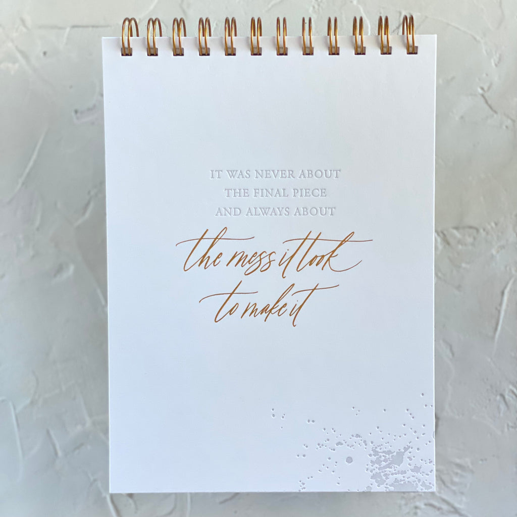 White notebook with white and gold text saying, “It Was Never About the Final Piece and Always About the Mess It Took to Make It”. Brass coil binding across the top.