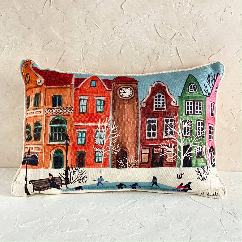 Pillow with image of a quaint small town in winter. Colorful houses all in a row with a clock tower in the center; white winter trees lining a skating pond in the middle; person sitting on a brown bench watching the skaters on the pond.