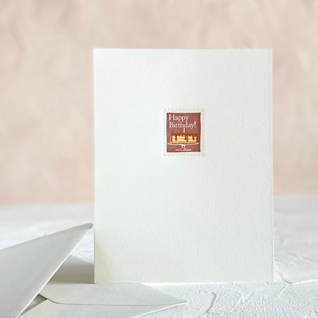 White card with image of a postage stamp with brown background and a vanilla cake with chocolate frosting. White text saying, “Happy Birthday”. A gray envelope is included.