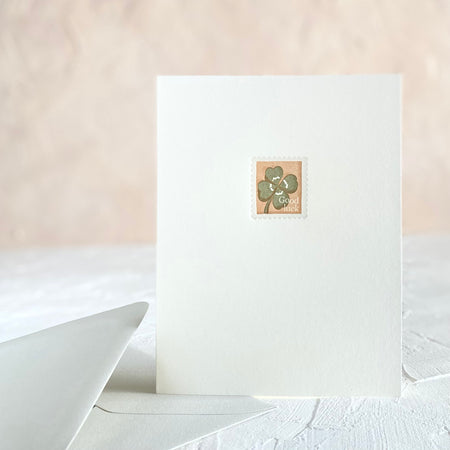 White card with image of a postage stamp with peach background and green four leaf clover. White text saying, “Good Luck” in bottom right corner.
