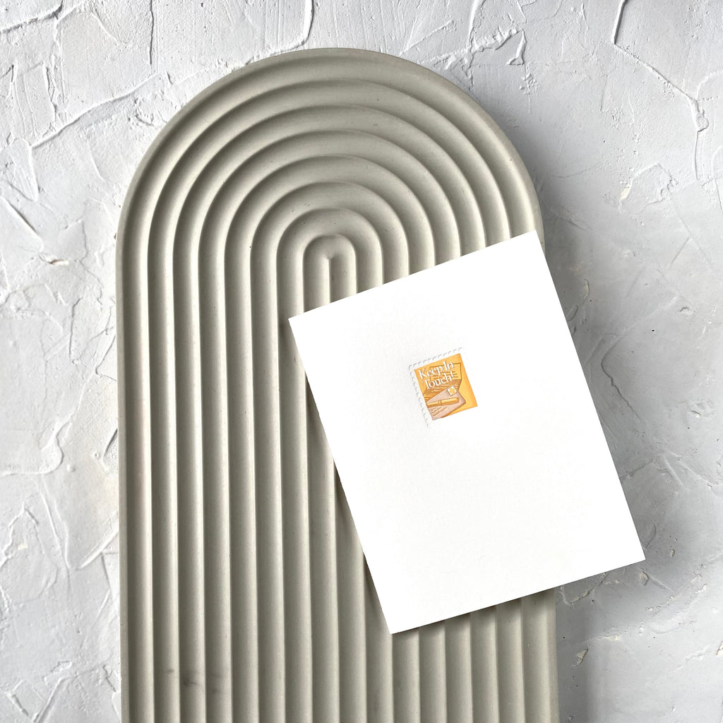 White card with image of a postage stamp with gold background with a letter, envelope and pen. White text saying, “Keep in Touch”. A gray envelope is included.