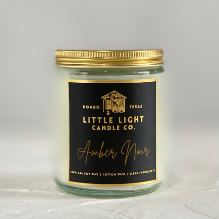 Glass jar with gold lid and black label with gold foil text saying, “Little Light Candle Co. Amber Noir”.