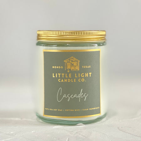 Glass jar with gold lid and gray label with gold foil text saying, “Little Light Candle Co. Cascades”. 