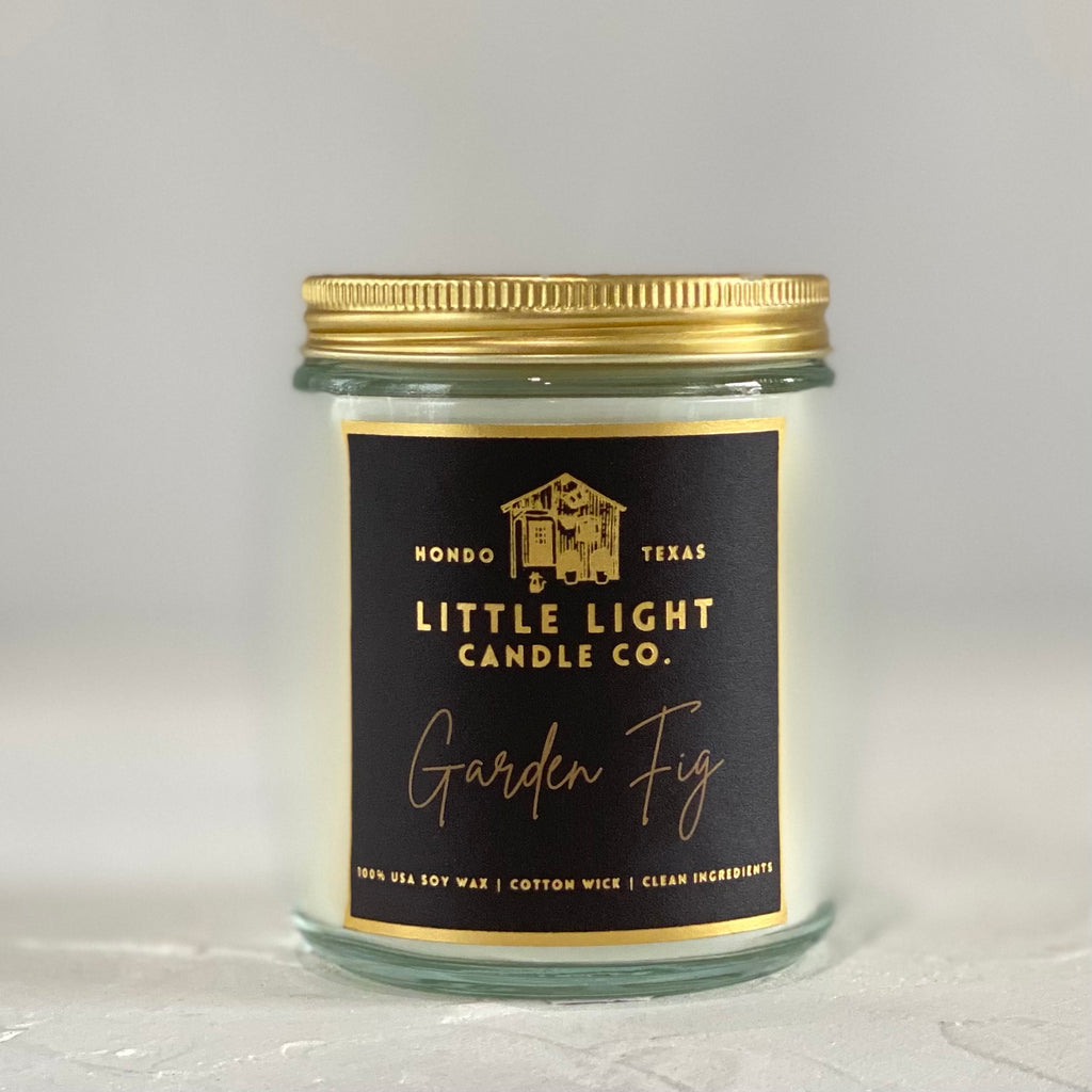 Glass jar with gold lid and black label with gold foil text saying, “Little Light Candle Co. Garden Fig”. 