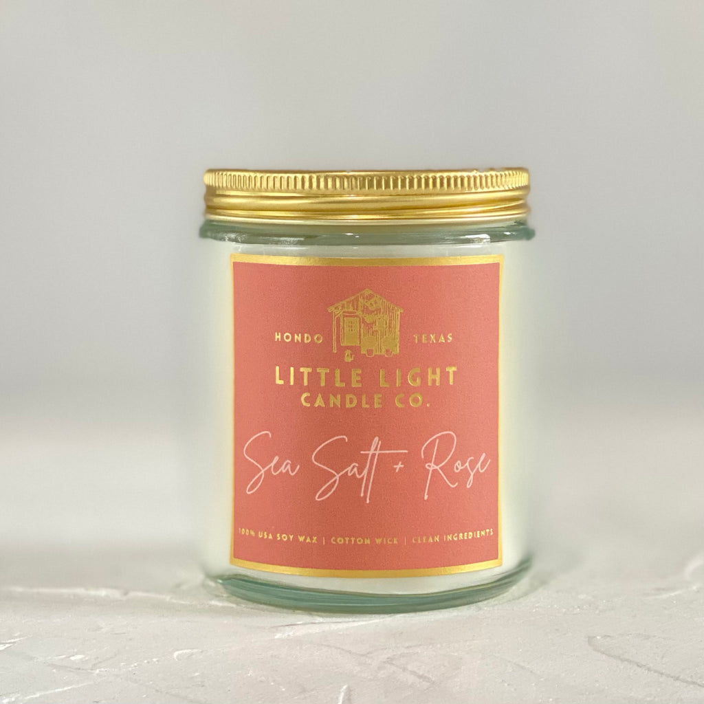 Glass jar with gold lid and orange label with gold foil text saying, “Little Light Candle Co. Sea Salt & Rose”.