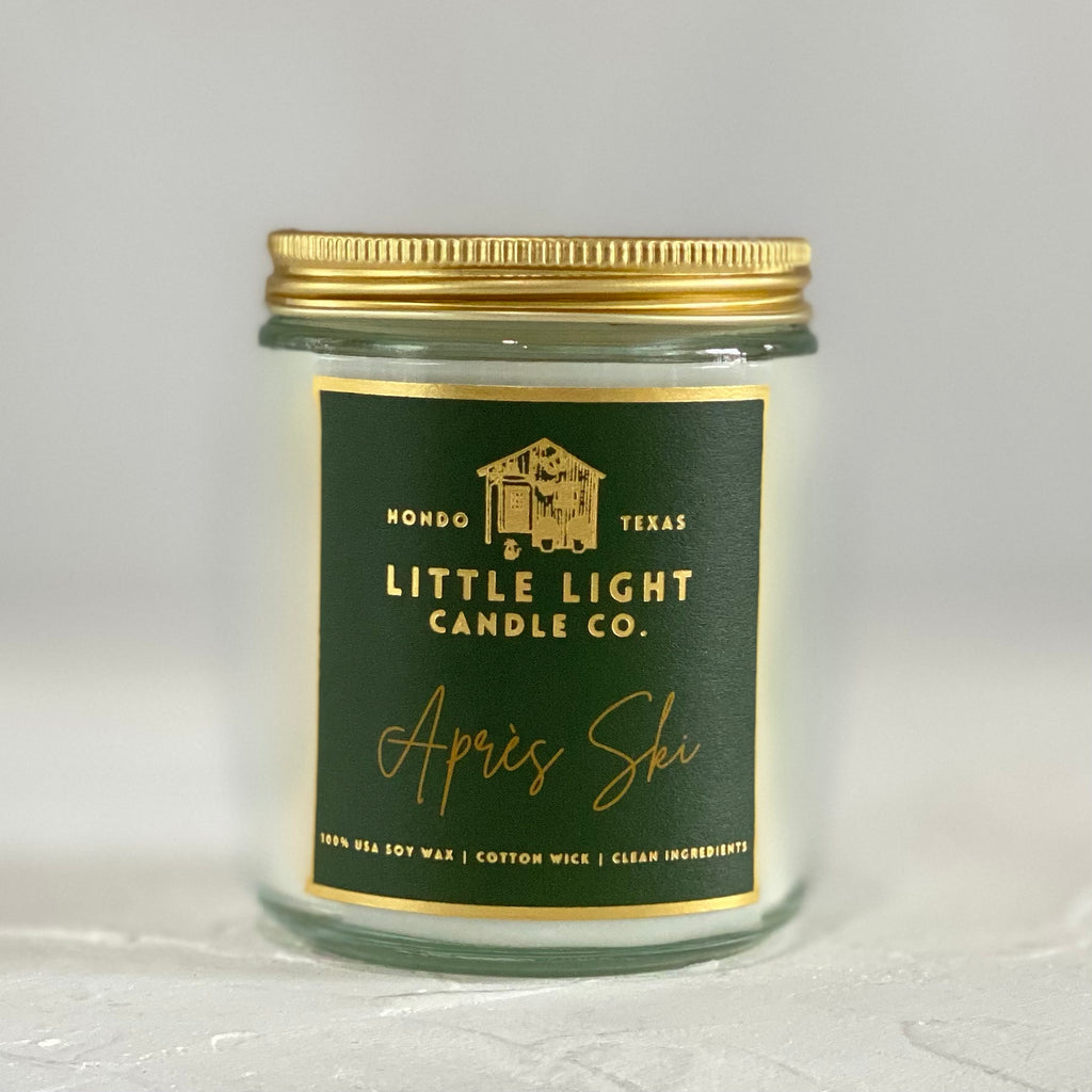 Glass jar with gold lid and green label with gold foil text saying, “Little Light Candle Co. Apres Ski”. 