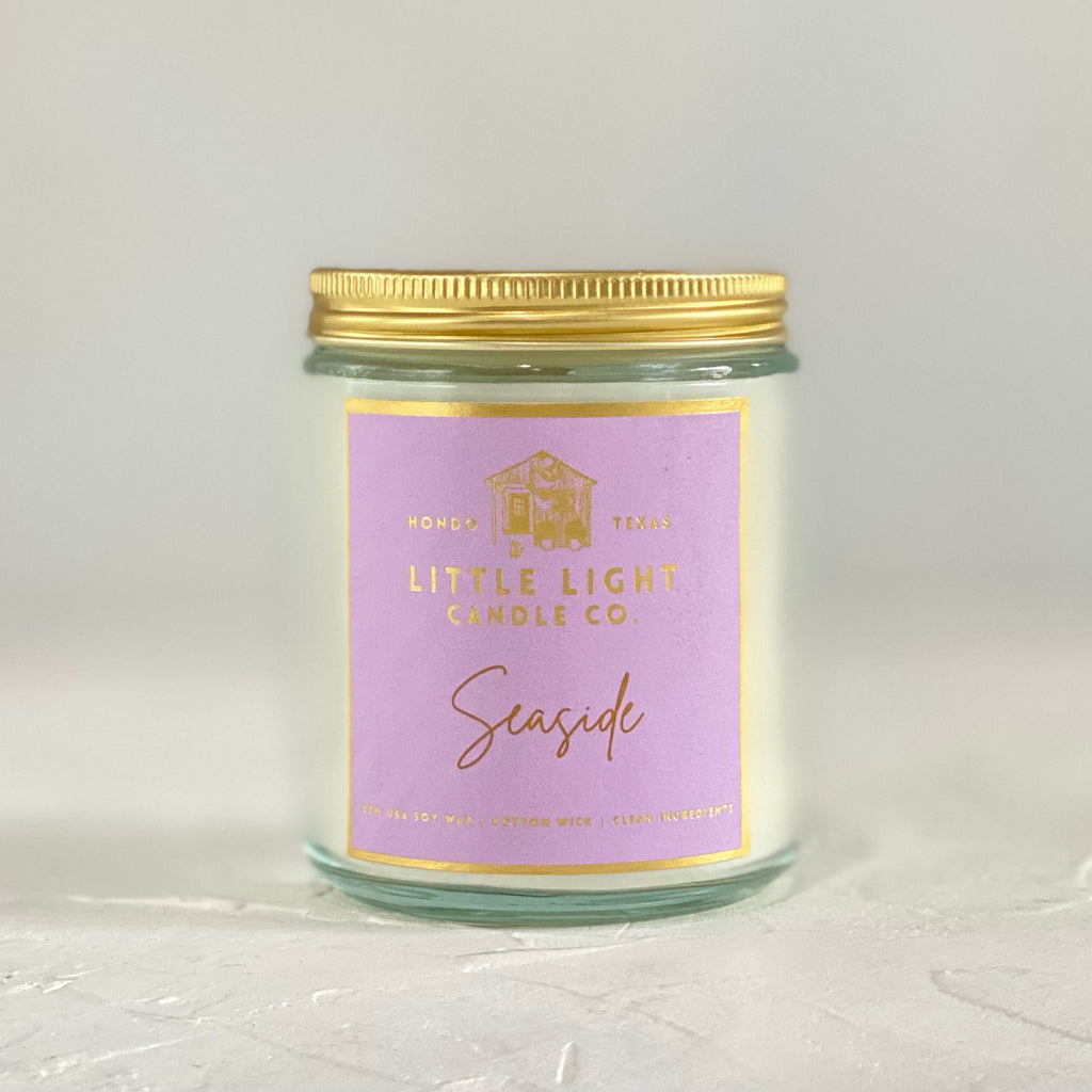 Glass jar with gold lid and purple label with gold foil text saying, “Little Light Candle Co. Seaside”.