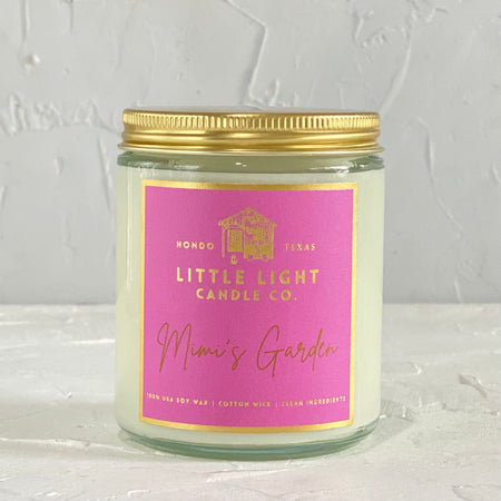 Glass jar with gold lid and pink label with gold foil text saying, “Little Light Candle Co. Mimi’s Garden”. 