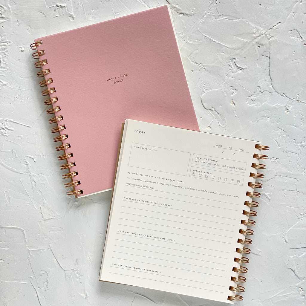 Notebook with dusty rose pink cover with gold foil text saying, “Daily Pause Journal”. Gold coil binding on left side.