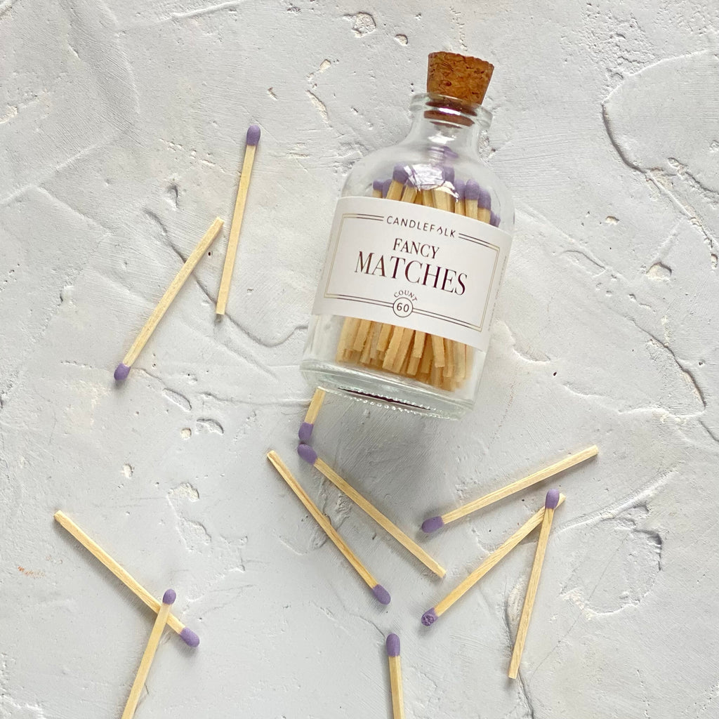 Small glass bottle with cork lid and white label with gold text saying, “Candlefolk Fancy Matches”. Filled with wooden matches with lavender tops.