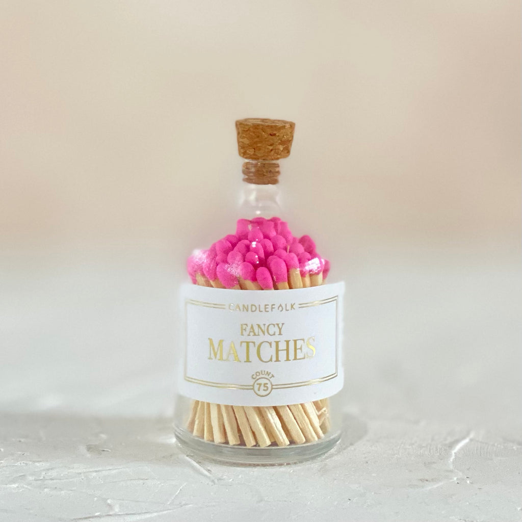 Small glass bottle with cork lid and white label with gold text saying, “Candlefolk Fancy Matches”. Filled with wooden matches with bright pink tops.