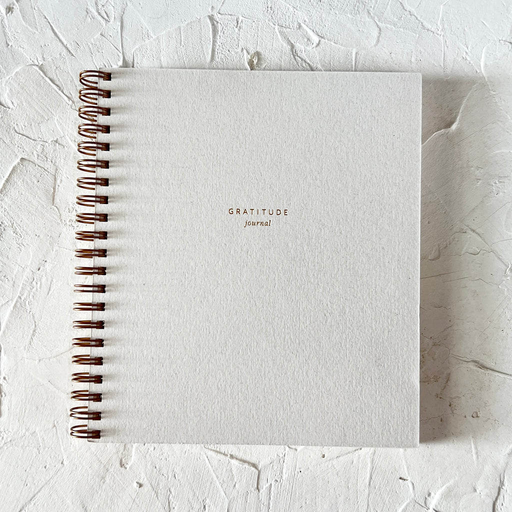 Notebook with white cover with gold foil text saying, “Gratitude Journal”. Gold coil binding on left side.