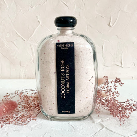 Glass bottle with black cap and black label with white text saying, “Native Nectar Coconut & Rose Floral Salt Soak”.