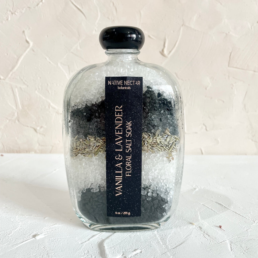 Glass bottle with black cap and black label with white text saying, “Native Nectar Vanilla & Lavender Floral Salt Soak”.