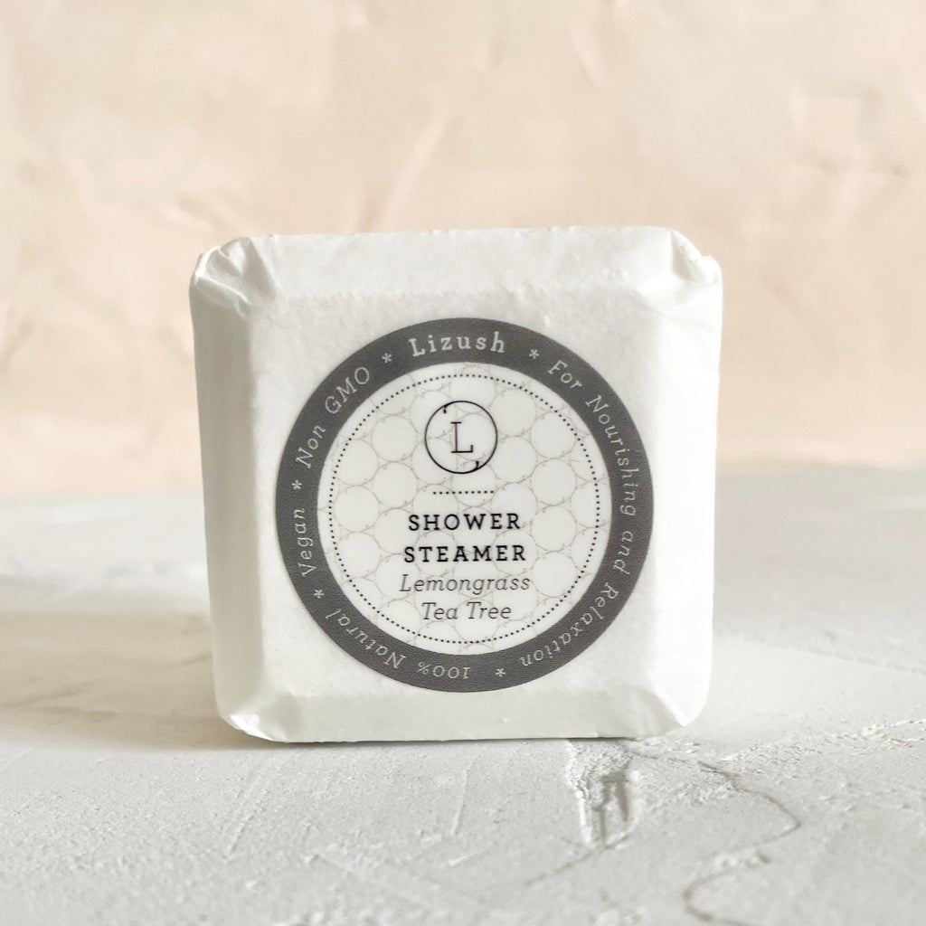 Wrapped in white square packaging with a gray circle in center. Black text saying, “Lizush Shower Steamer Lemongrass and Tea Tree.”