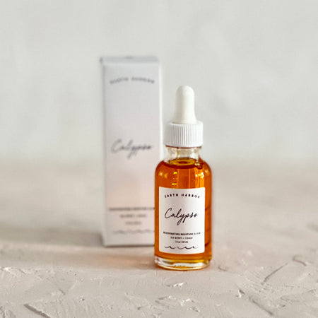 Clear glass bottle with white dropper top. White label with black text saying, “Earth Harbor Calypso Facial Oil”. Liquid is orange color. Packaged in a white box.