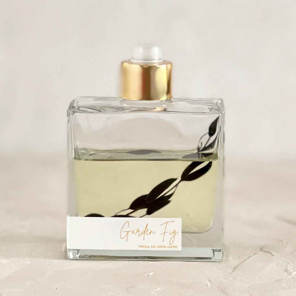Square glass bottle with gold band spritzer and white label with gold foil text saying, “Garden Fig Freesia, Fig, Green Leaves”.