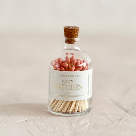 Small glass bottle with cork lid and white label with gold text saying, “Candlefolk Fancy Matches”. Filled with wooden matches with coral tops.