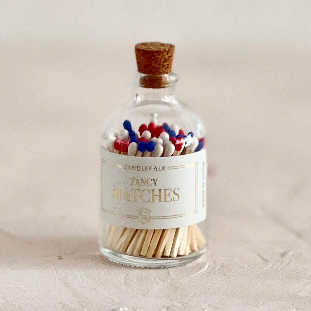 Small glass bottle with cork lid and white label with gold text saying, “Candlefolk Fancy Matches”. Filled with wooden matches with red, white and blue tops.