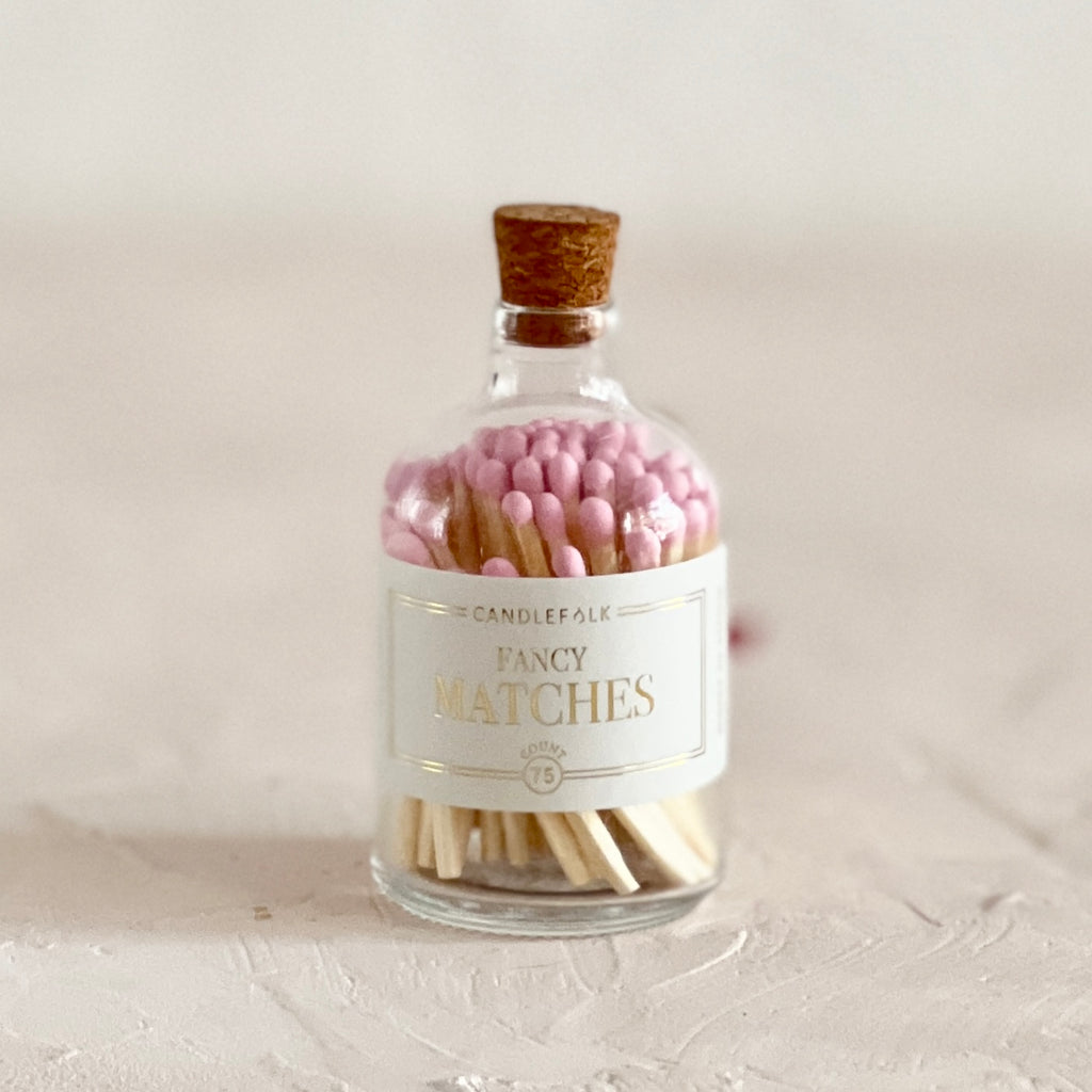 Small glass bottle with cork lid and white label with gold text saying, “Candlefolk Fancy Matches”. Filled with wooden matches with light pink tops.