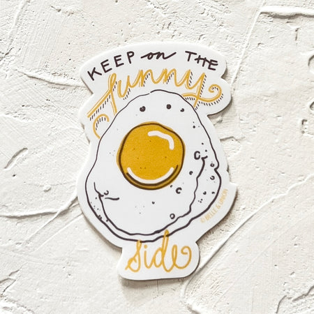 White sticker with gray and yellow text saying, “Keep On the Sunny Side”. Image of a sunny side up egg with a smiley face. 