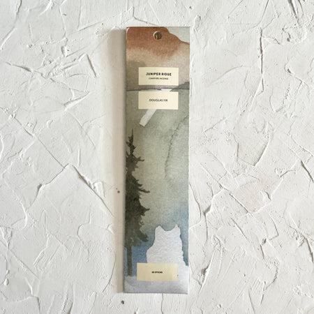 White vertical rectangle packaging with image of a fir tree and muted mountains. Black text in an ivory box saying, “Juniper Ridge Campfire Incense Douglas Fir”.